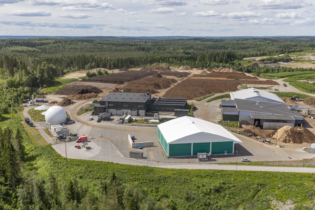 Ilmakuva metsän ympäröimästä biokaasu- ja kompostointilaitoksesta, jonka alueella näkyy tuotantorakennuksia, kaasusäiliö sekä suuria kompostiaumoja.