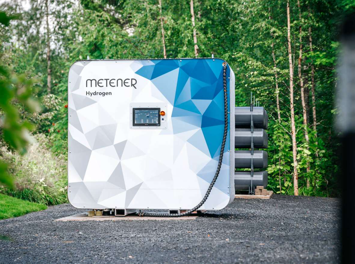 White-and-blue Metener hydrogen electrolyser installed outdoors, with stacked cylinders beside it and trees in the background.